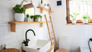 Interior of small bathroom with window, wooden decoration, shelf and ladder. Stylish wash basin and tap, round mirror on a white wall and plants on a shelf. Rustic bathroom in a cottage in rural style