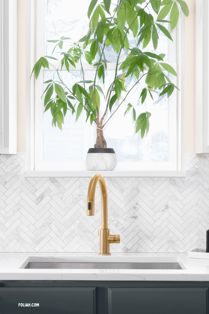 13 Gorgeous Kitchen Plant Styling Ideas You’ll Want to Copy 7 at Foliah Modern kitchen sink area with a gold faucet, white marble countertop, and herringbone tile backsplash, featuring a money tree plant on the windowsill above.