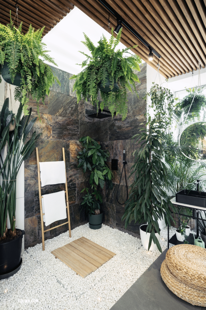 Shower area in a modern bathroom with hanging ferns, bird of paradise, and dracaena, styled with stone tiles, pebble flooring, and a bamboo towel ladder for a jungle-inspired look.