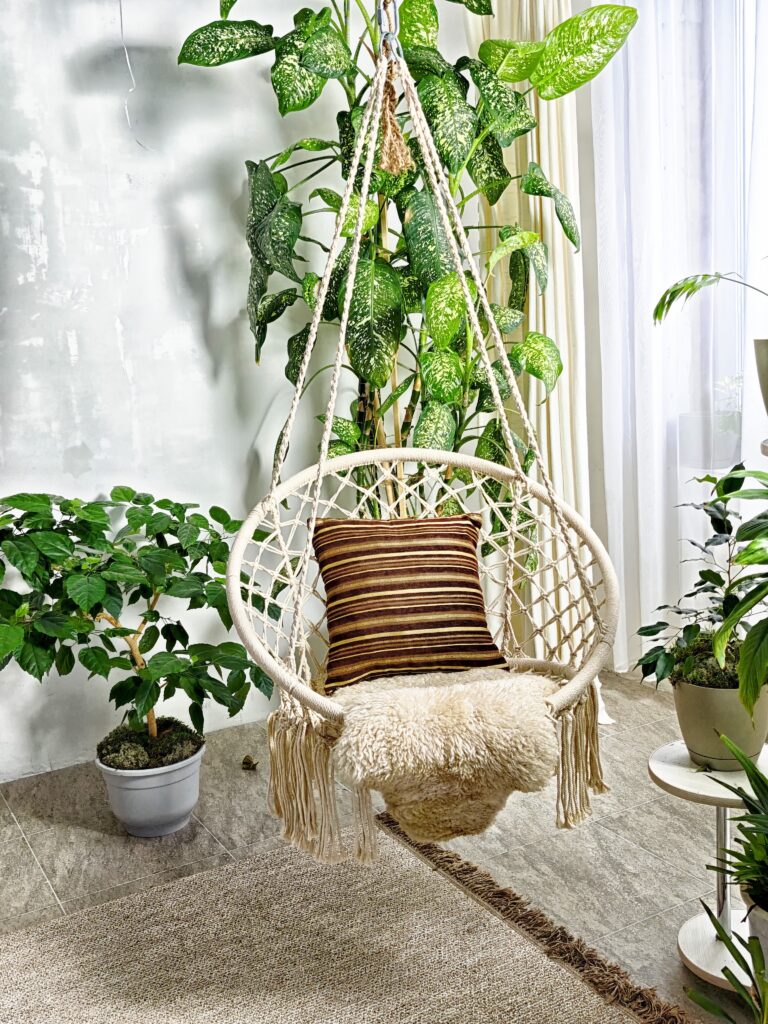 A wicker hanging swing chair with a soft pillow is displayed in a bright room filled with houseplants and boho style decor.